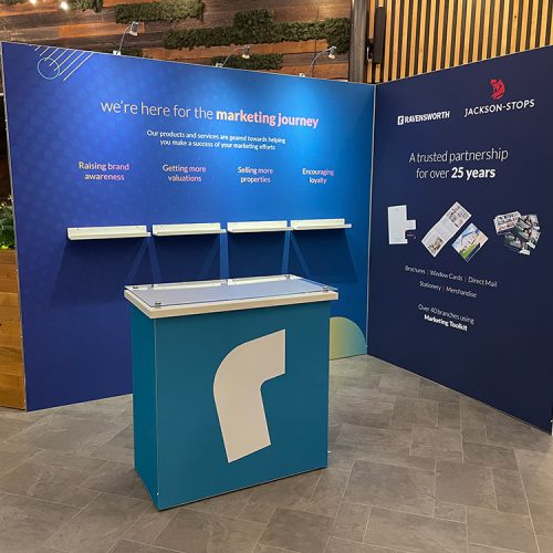 Modular tension fabric exhibition stand with magnetic shelving for literature display, blue graphic panels and a small reception desk in a hotel foyer