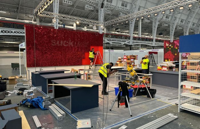 the build of a custom modular stand for suck uk at top drawer in olympia, with system back wall, and desks and custom elements like the red shimmer wall