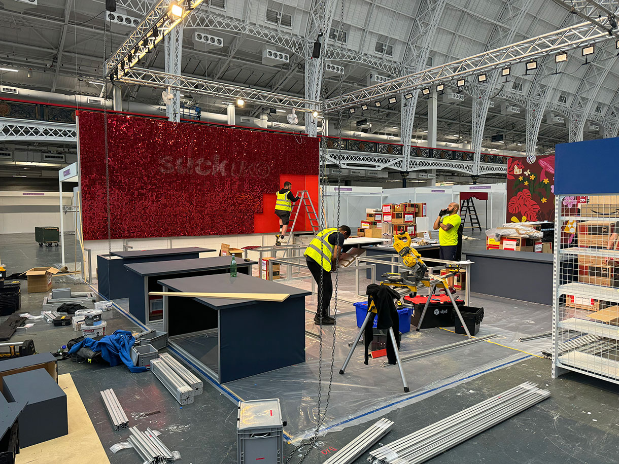 the build of a custom modular stand for suck uk at top drawer in olympia, with system back wall, and desks and custom elements like the red shimmer wall